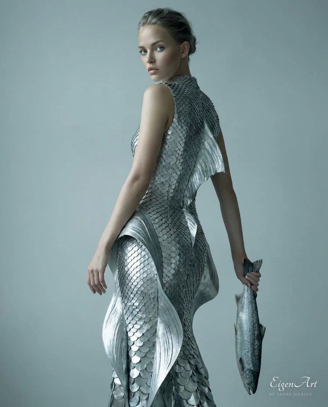 Model in silberner Schuppen-Robe von hinten, einen Fisch wie eine Clutch haltend